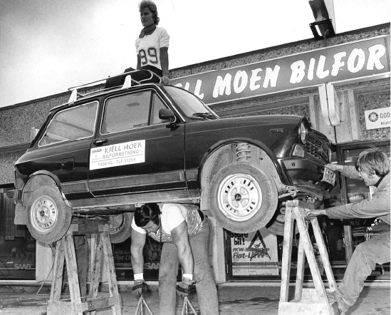 Tre personer jobber med en liten bil på trestativ foran en bygning.