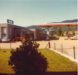 Saab-forhandler og bensinstasjon med bil, traktor og busk foran.