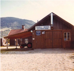 Trehus med skilt "Kjell Moen Bilforretning", rød traktor utenfor.