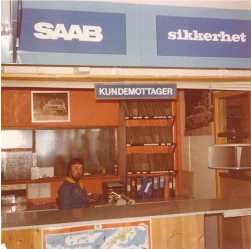 Person bak skranke under "SAAB", "sikkerhet", "KUNDEMOTTAKER"-skilt.